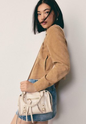 Mladá žena s čiernymi vlasmi má na sebe béžový semišový kabát a džínsovú sukňu, drží malú krémovú kabelku na jednoduchom pozadí.