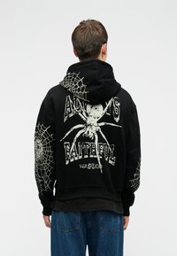 Cilvēks, kas ģērbts melnā kapuces džemperī ar baltām zirneklēm un tīkla dizainiem, un ar tekstu "Vienmēr uzticīgs", kopā ar zilu džinsu biksēm, stāv ar muguru.