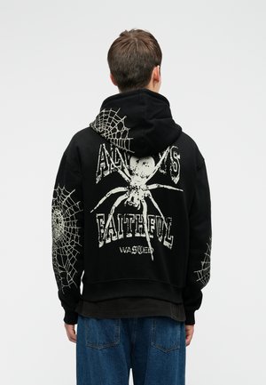 Cilvēks, kas ģērbts melnā kapuces džemperī ar baltām zirneklēm un tīkla dizainiem, un ar tekstu "Vienmēr uzticīgs", kopā ar zilu džinsu biksēm, stāv ar muguru.