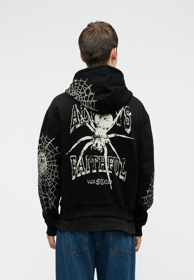 Cilvēks, kas ģērbts melnā kapuces džemperī ar baltām zirneklēm un tīkla dizainiem, un ar tekstu "Vienmēr uzticīgs", kopā ar zilu džinsu biksēm, stāv ar muguru.