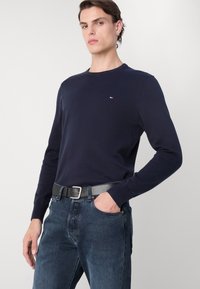 Marineblauer Pullover mit langen Ärmeln und Rundhalsausschnitt, mit gerippten Bündchen, versehen mit einem kleinen Logo auf der Brust, kombiniert mit dunkelblauen Jeans und einem grauen Gürtel.