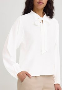 Chemisier blanc avec un col rond, des manches bouffantes longues et une fermeture à nouer à l'avant. Fabriqué en tissu léger avec une surface légèrement texturée.