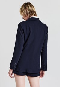 Blazer à rayures navy avec manches longues et revers crantés ; comporte cinq boutons aux poignets et dispose d'une coupe légèrement cintrée.