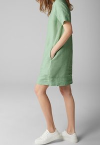 Grünes Leinenkleid mit kurzen Ärmeln, seitlichen Taschen und lockerem Schnitt, kombiniert mit weißen Sneakers. Glatte Textur, Midi-Länge bis zur Oberschenke.