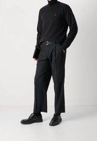 Černý rolák se logem, černé kalhoty na míru a černé kožené boty. Drží černé zařízení, celkový elegantní monochromatický vzhled.