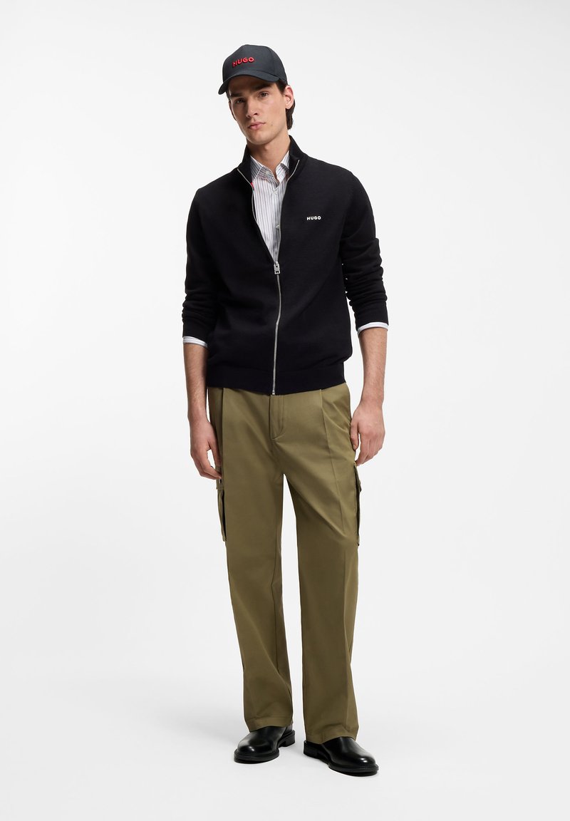 Jeune homme portant une veste noire zippée, une chemise rayée, un pantalon cargo vert olive, des chaussures en cuir noir et une casquette noire avec un texte rouge.