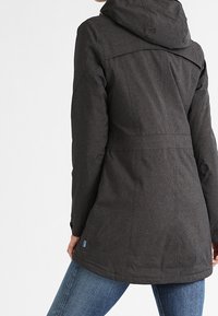 Veste longue gris foncé avec une capuche. Présente un dos structuré et un tissu texturé. Associée à un jean en denim bleu.