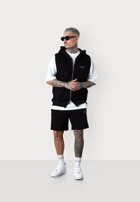 Schwarze Zip-Jacke über einem weißen T-Shirt, schwarze Shorts, weiße Sneakers und Sonnenbrille; zeigt ausgefallene Tattoos auf den Armen.