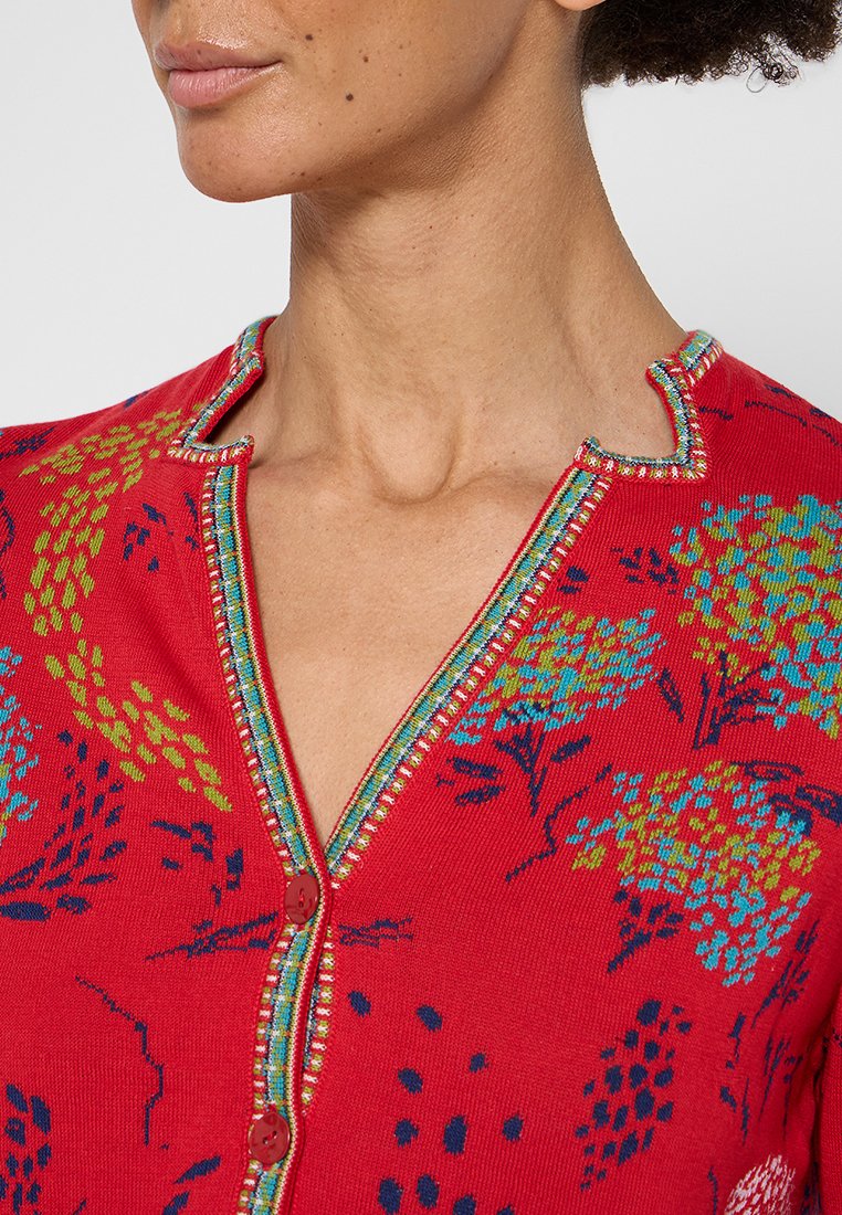Czerwony sweter z dzianiny z niebieskimi i zielonymi wzorami kwiatowymi. Posiada teksturowany kołnierz z wielokolorowym wykończeniem oraz zapięcie na guzik.