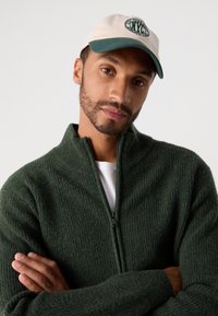 Mann med trimmet skjegg som har på seg en grønn zip-up-genser og en hvit og grønn NYC baseball caps, med armene krysset mot en enkel bakgrunn.