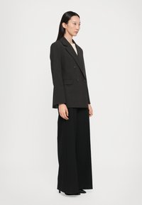 Blazer doppiopetto grigio scuro con rever a punta, taschino singolo sul petto e vestibilità sartoriale, abbinato a pantaloni neri a gamba larga.