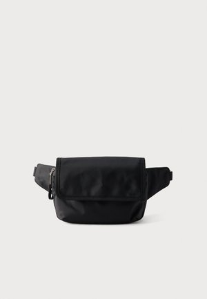 Sac banane en nylon noir avec rabat frontal et poche latérale zippée, sangle réglable, sur un fond clair uni.