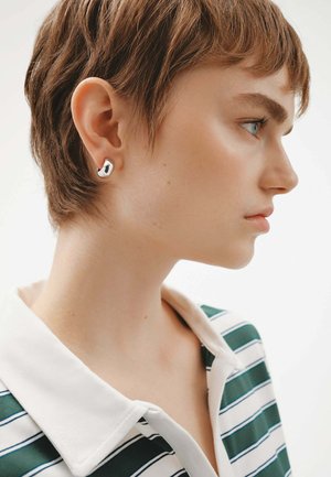 Pendiente de plata con forma redonda, situado en la oreja izquierda. El modelo lleva una camisa con cuello de rayas verdes y blancas.