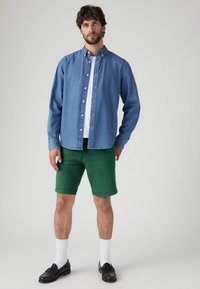 Lys blå denim button-up skjorte, hvid t-shirt, grøn shorts, sorte loafers, hvide sokker, står på en neutral baggrund.