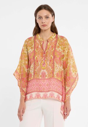 Vrouw die een losse, luchtige blouse draagt met roze en gele ingewikkelde paisley- en bloemenpatronen, gecombineerd met een witte broek, staand tegen een witte achtergrond.