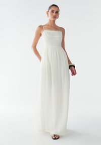 Vestido maxi blanco con tirantes finos, tejido texturizado y borde de encaje circular alrededor del cuerpo y el dobladillo. Modelo posa con las manos en los bolsillos.