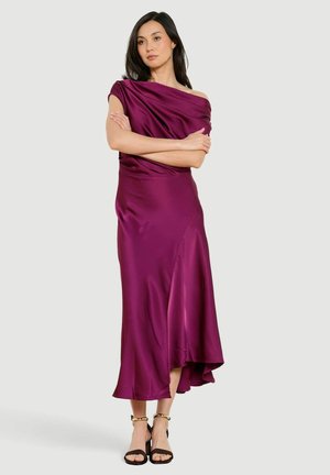 Woman standing with arms crossed wearing a sleeveless, off-shoulder, asymmetrical purple satin dress and black heeled sandals.