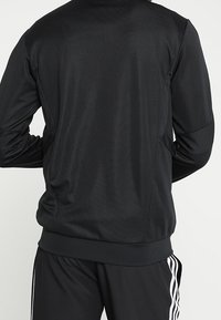 Svart idrottsjacka med en jämn textur, figurnära design och ribbade manschetter. Visad på en modell som bär matchande svarta byxor.