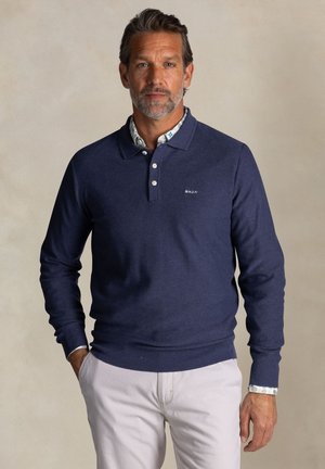 Marineblauwe polo met lange mouwen, gemaakt van gestructureerde stof, met een knoopsluiting en geribbelde manchetten, gedragen over een bloemenpatroon shirt.