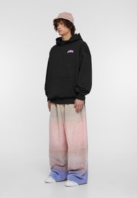 Juodas džemperis su priekiu kišene ir rožiniu logotipu, derinamas su plačiomis pastelinėmis kelnių su gradientu rožinės ir mėlynos spalvų, užbaigtas baltais sportbačiais.