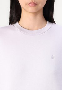 Sweatshirt de culoare lavandă deschisă, cu un decolteu rotund cu nervuri, având un mic logo brodat pe pieptul stâng. Textura materialului este netedă.