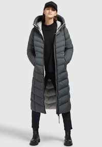 Un long manteau rembourré gris foncé avec un motif matelassé horizontal, une capuche surdimensionnée, une fermeture éclair sur le devant et des poches ; porté par-dessus un pull noir et un pantalon.