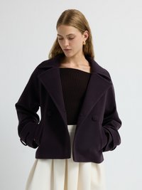 Cappotto corto di colore viola scuro con ampio colletto a scollo a V, bottoni doppi e tasche laterali. Tessuto morbido e strutturato.