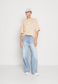 Beige oversized t-shirt med fronttryk, lyseblå brede jeans, hvide slip-on sko, og en lys grå cap med et sort logo.