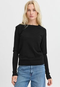 Maglione lungo nero con scollo a bardot, realizzato in un tessuto a maglia morbido. Polsini e orlo a coste, abbinato a jeans di denim blu chiaro.