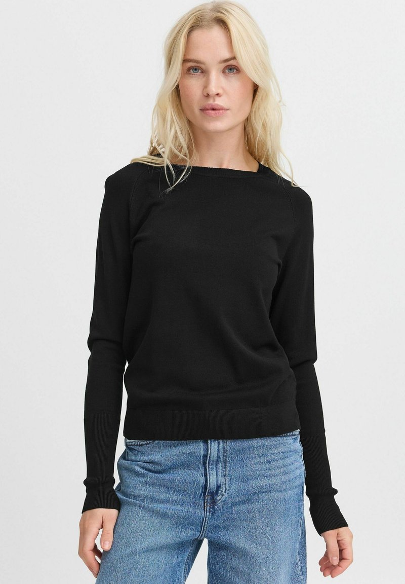 Maglione lungo nero con scollo a bardot, realizzato in un tessuto a maglia morbido. Polsini e orlo a coste, abbinato a jeans di denim blu chiaro.