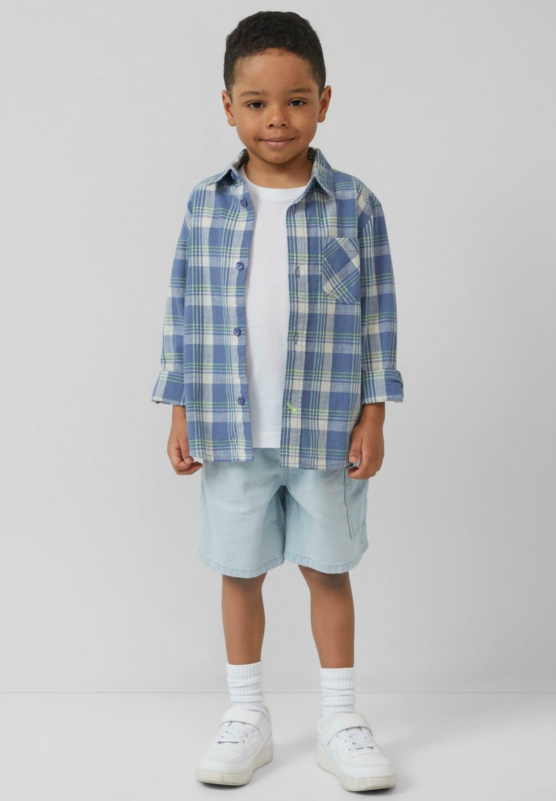 Blauw en groen geblokt overhemd met knopen over een wit T-shirt, gecombineerd met lichtblauwe shorts en witte sneakers met geribde enkelsokken.