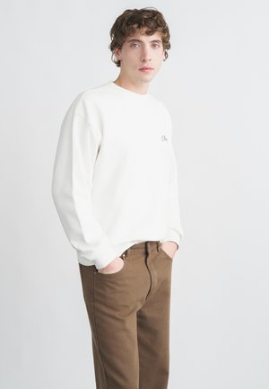 Hvid sweatshirt med løs pasform og ribstrikkede manchetter, kombineret med brune bukser. Sweatshirten har en lille sort broderet detalje.