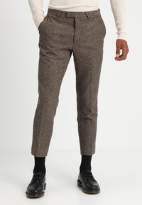Brown herringbone trousers with tapered legs, front pockets, and a flat front design. Paired with black shoes and dark socks.