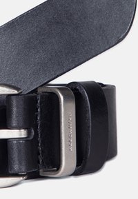 Cinturón de cuero negro con un acabado suave, que presenta una hebilla de metal plateado y una correa ajustable. Detalle de logotipo grabado en la hebilla.