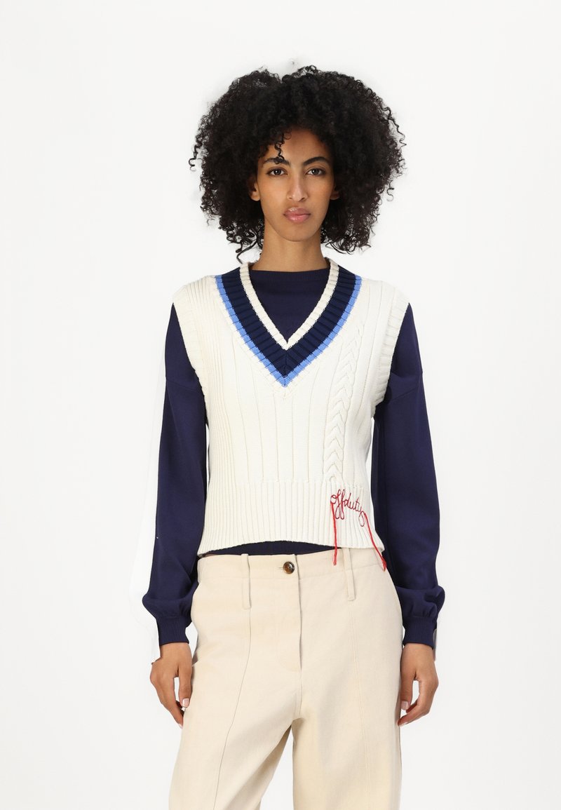 White knitted sleeveless sweater with V-neck and blue trim, featuring cable knit texture and red embroidered detail, worn over a navy long-sleeve shirt.