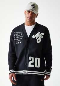 Cardigan noir en tricot avec des accents blancs, arborant un logo "M", un patch "20" et un texte brodé. Il a un col en V et des poignets rayés.