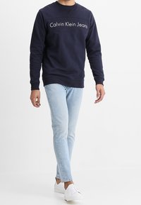 Felpa blu navy con logo "Calvin Klein Jeans", scollo rotondo, maniche inserite, indossata con jeans azzurri e sneakers bianche.