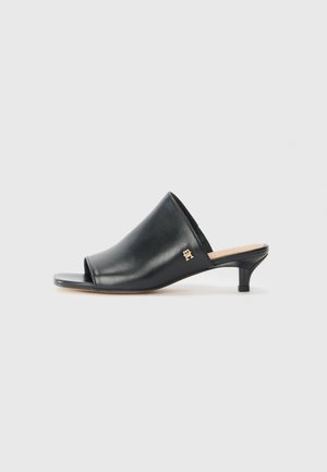 Mule de cuero negro con abertura en la punta, textura suave, tacón bajo y detalle de logo en tono dorado. Diseño sencillo con una silueta estilizada.