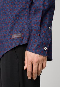 Cămașă cu mânecă lungă de culoare bleumarin, cu un model geometric roșu și albastru. Prezintă un etichetă, manșete cu nasturi și o textură a materialului neted.