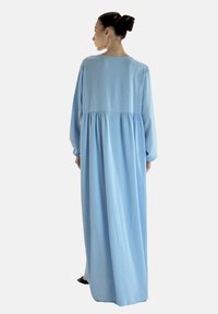 Długa, zwiewna, jasnoniebieska sukienka z miękkiego materiału, z długimi rękawami i marszczoną talią, sięgająca aż do kostek.