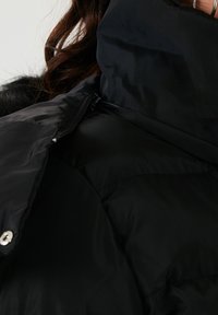 Chaqueta de plumas negra con acabado brillante, con cierre frontal de cremallera, gran cuello y botones de presión plateados. Textura suave con patrón acolchado.