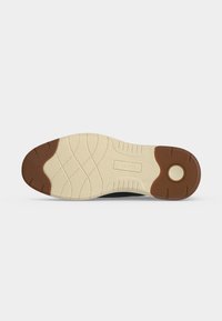 Semelle de chaussure avec des sections en caoutchouc beige et marron, un motif de bande de roulement et une zone circulaire marron près du talon sur fond blanc.