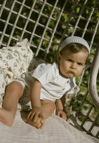 Ljust beige kortärmad body med knapprad, liggande på en texturerad filt med en blommönstrad kudde, i en vävd stol.