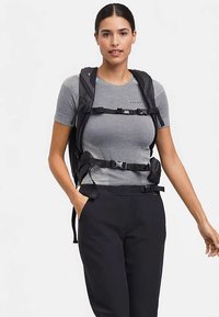 Zwarte rugzak met verstelbare riemen, gespen en een gestroomlijnd ontwerp, gedragen over een grijs shirt met korte mouwen en donkere broek.