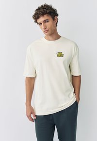 Vaaleanbeige puuvillainen t-paita, jossa on lyhyet hihat ja vasemmassa rintakehässä värikäs "Outkast"-logo. Pehmeä materiaali.