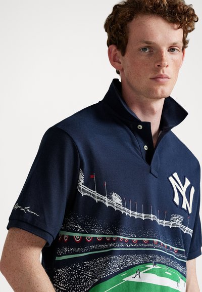 Νέος άνδρας με ανοιχτόχρωμο δέρμα και κόκκινα σγουρά μαλλιά, φορώντας ναυτικό μπλουζάκι πόλο με λογότυπο των New York Yankees και γραφικό σχέδιο γηπέδου μπέιζμπολ.