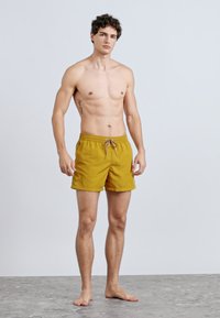 Sárga fürdőnadrág rugalmas derékpánttal és húzózsinórral, könnyű anyagból készült. Laza szabású és egyszínű dizájnú.