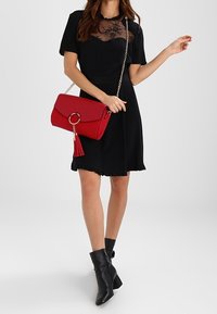 Abito nero con dettagli in pizzo, borsa rossa con anello e nappina, e stivali neri alla caviglia con tacco largo, che mettono in risalto elementi di design testurizzati.