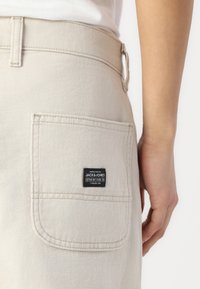 Beige denim pants with a back pocket featuring a black label. The fabric has a smooth texture and visible stitching details.