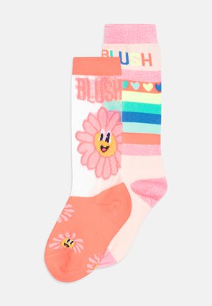 Deux paires de chaussettes : une blanche avec des fleurs roses et un smiley, et une pêche avec des rayures et le texte "BLUSH" dans un mélange de couleurs. Matière en mélange de coton.